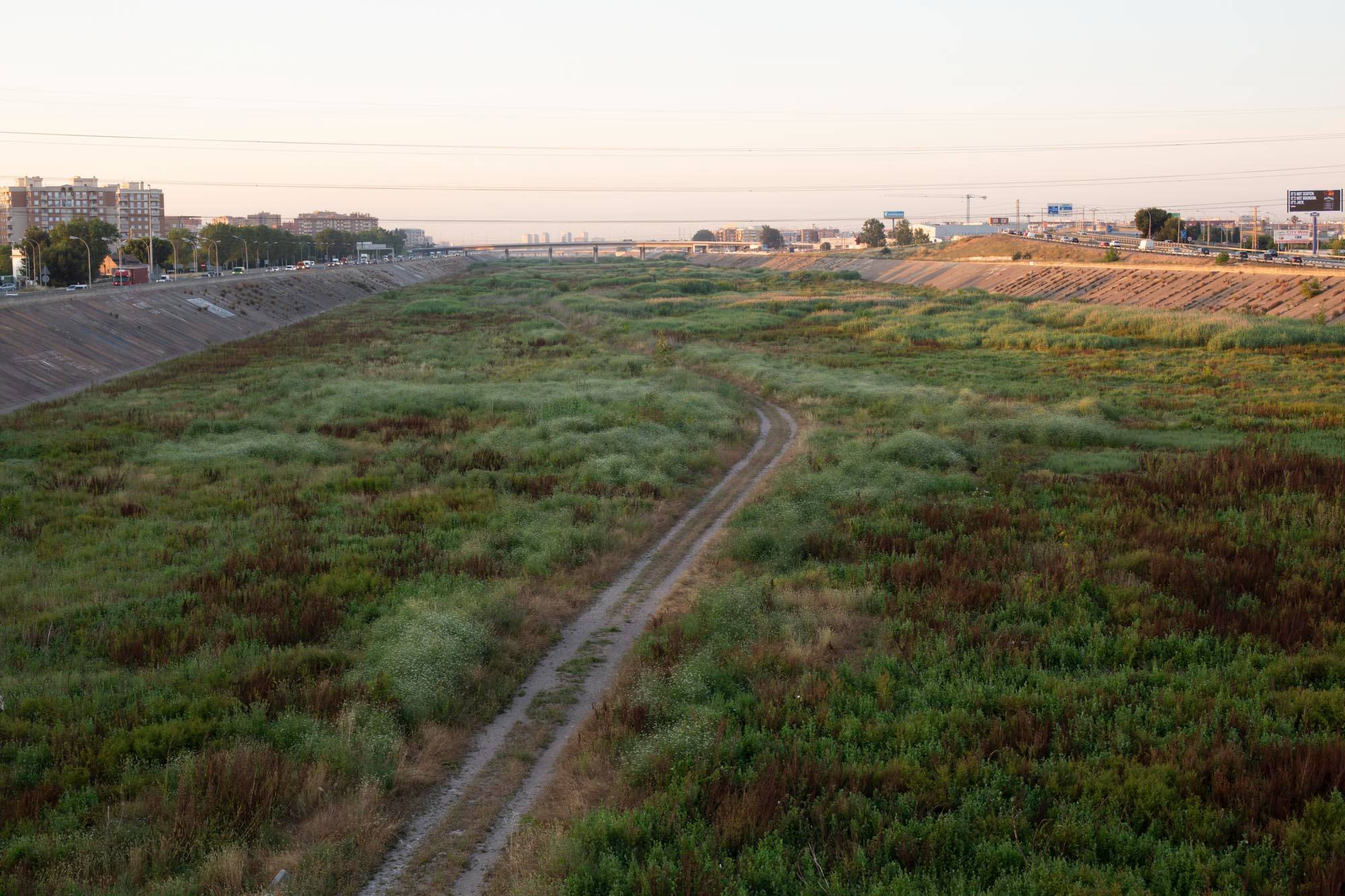 Color photograph of a grassy patch with buildings in the distance, two track road leads towards the buildings.